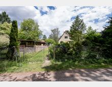 Blick auf ein eingezäuntes Wohngrundstück mit Metalltor und schmalem Weg, umgeben von dichtem Grün und hohen Bäumen. Im Hintergrund stehen ein kleines Haus mit steilem Giebeldach sowie ein niedriger Anbau mit Natursteinmauer und Flachdach.