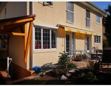 Zweistöckiges Wohnhaus mit beiger Fassade und großen Fenstern mit weißen Rahmen. Eine gelbe Markise spendet Schatten über einer Terrasse, die mit Gartenmöbeln und Pflanzen dekoriert ist; ein kleiner Gartenbereich befindet sich im Vordergrund.
