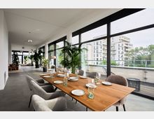 Modernes Esszimmer in einem lichtdurchfluteten Raum mit deckenhohen Glasfenstern, die den Blick auf eine Terrasse mit Stadtgebäuden freigeben. Holz-Esstisch mit eingedeckten Tellern und Gläsern, umgeben von gepolsterten Stühlen; zahlreiche Pflanzen und gemütliche Sitzbereiche im Hintergrund.