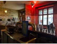 Innenansicht einer Bar mit Tresen aus Edelstahl, Gläsern und Barwerkzeugen im Vordergrund. Die Wände sind dicht mit Graffiti bedeckt, rotes Licht und eine Discokugel hängen von der Decke, daneben große Fenster zur Straße.