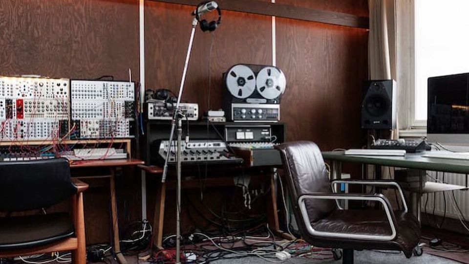 Aufnahme eines Tonstudios mit holzvertäfelter Wand, Misch- und Synthesizer-Modulen, Bandmaschine sowie Mikrofonständer. Rechts stehen ein Schreibtisch mit Monitor und Lautsprecher neben einem großen Fenster mit Vorhängen; auf dem Boden liegen zahlreiche Kabel.
