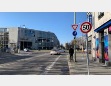Straßenszene in einer Stadt mit einem modernen Bürogebäude im Hintergrund, beschriftet mit „Aufbau Haus“ und „Planet Modulor“. Im Vordergrund stehen Verkehrsschilder, darunter ein „50 km/h“-Schild, sowie Bürgersteige, eine Straße und parkende Fahrräder.
