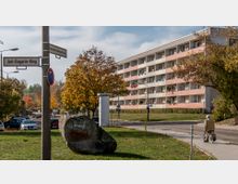 Mehrstöckiges Wohngebäude mit Balkonen und weiß-rosa Fassadenelementen, umgeben von Bäumen mit herbstlichem Laub. Im Vordergrund eine Straßenecke mit Straßenschildern und einem großen Stein mit Graffiti, sowie ein Fußgängerweg, auf dem eine ältere Person läuft.