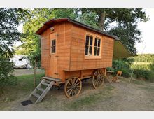 Holzbauwagen auf großen hölzernen Rädern, mit einer kleinen Holztreppe und einem Fenster an der Seite, auf einer Wiese nahe Bäumen und einem Campingplatz. Daneben stehen ein kleiner Holztisch und zwei Stühle unter einem Sonnensegel.