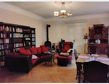 Wohnzimmer mit klassischem Interieur: Ein großer Bücherregal an der linken Wand, ein dunkelrotes Sofa mit roten Kissen, ein schwarzer Kaminofen in der Ecke neben einer weißen Doppeltür, ein massiver Holzesstisch mit Stühlen rechts im Vordergrund und ein antikes Sideboard im Hintergrund. Der Holzboden ist in Fischgrätenmuster verlegt, und eine zentrale Deckenlampe beleuchtet den Raum.