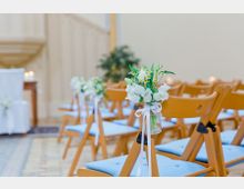 Holzstühle mit hellblauen Sitzpolstern in Reihen angeordnet, dekoriert mit kleinen Blumensträußen aus weißen Blüten und Schleifen an den Lehnen, in einem hellen, schlichten Raum mit Kerzen und Pflanzen im Hintergrund.