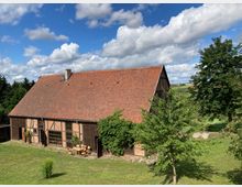 Fachwerkhaus mit roten Ziegeln und großen Fenstern, umgeben von einer grünen Wiese und einzelnen Bäumen. Im Vordergrund steht ein Holztisch mit Stühlen, im Hintergrund eine ländliche Landschaft mit Feldern und Bäumen unter blauem Himmel mit Wolken.