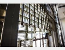 Nahaufnahme eines Fensters mit Glasbausteinen und vertikalen Metallgittern, die das Fenster abdecken. Die Umgebung wirkt industriell mit sichtbaren Stahl- und Betonstrukturen.