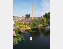 Industriegebäude an einem Flussufer mit einem hohen Backsteinschornstein, umgeben von grüner Vegetation und Seerosen im Wasser. Eine gelbe Boje schwimmt auf der Wasseroberfläche, der Himmel ist teils bewölkt.