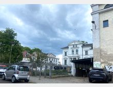 Straßenszene mit einer weißen, historischen Villa mit Säulenelementen und Rundbogenfenstern im Hintergrund. Rechts im Vordergrund ist ein altes Gebäude mit abgeplatztem Putz sowie ein Tor zu sehen, links parkende Autos und Bäume unter bewölktem Himmel.