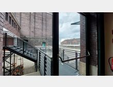 Blick durch eine Glastür auf eine Außentreppe mit Metallgeländern in einem Industriegebäude aus Backstein. Im Hintergrund befinden sich weitere Backsteingebäude und eine gepflasterte Fläche unter bewölktem Himmel.