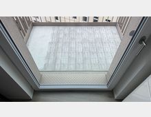 Blick durch eine Glastür auf einen kleinen Balkon mit hellgrauem Betonboden und umlaufendem Metallgeländer. Im Hintergrund sind die Fassaden und Fenster eines benachbarten Gebäudes zu sehen.