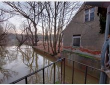 Eine ruhige Flusslandschaft mit kahlen Bäumen am Ufer im Winter, rechts ein altes, verwittertes Gebäude mit einem teils freigelegten Ziegelmauerwerk. Im Vordergrund befindet sich ein Geländer, das einen kleinen Balkon oder Steg andeutet.