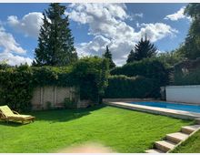 Garten mit großer Rasenfläche, einer Sonnenliege, einem rechteckigen Pool mit Steinumrandung und einer begrünten Mauer, die von dichten Kletterpflanzen und Bäumen umgeben ist. Im Hintergrund sind hohe Nadelbäume und ein bewölkter Himmel zu sehen.