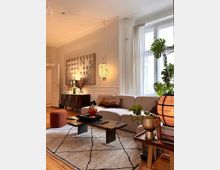 Helles Wohnzimmer mit einer cremefarbenen Couch, einem modernen Couchtisch aus dunklem Holz und einem gemusterten Teppich. Ein großes Fenster mit weißen Vorhängen lässt natürliches Licht herein, ergänzt durch eine hängende Lampe und dekorative Pflanzen.