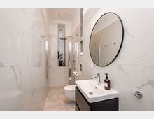 Schmales, modernes Badezimmer mit großformatigen, marmorierten Wandfliesen, bodengleicher Glasdusche mit Regendusche und einem Waschbecken auf dunklem Unterschrank. Ein runder Wandspiegel, ein wandhängendes WC und ein hohes Fenster am Ende des Raums ergänzen die helle, minimalistische Ausstattung.