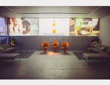 Moderner Lounge-Bereich mit schlichten Möbeln, darunter orangenförmige Stühle in der Mitte und gepolsterte Sitzbänke an den Seiten. Im Hintergrund wird eine großformatige Projektion mit Getränkewerbung auf eine Betonwand gezeigt.