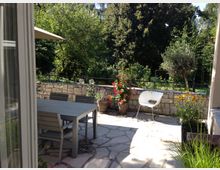 Eine gepflasterte Terrasse mit einem grauen Tisch und Stühlen, flankiert von Blumentöpfen mit bunten Pflanzen. Im Hintergrund sind eine niedrige Steinmauer und üppige grüne Bäume zu sehen.