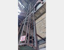 Eine industrielle Innenansicht mit einer schmalen, rostigen Metallleiter, die von einem Schutzkäfig umgeben ist. Die Umgebung zeigt Backsteinwände, Stahlträger und ein Schild am unteren Leiterbereich mit einem Verbotssymbol für den Zugang.