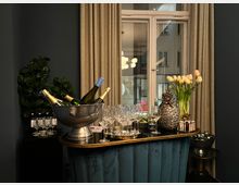 Barbereich in einem elegant gestalteten Raum mit einem kleinen, gepolsterten Tresen in Blau. Auf der Bar stehen ein Getränkekühler mit Weinflaschen, Gläser, eine dekorative Ananas aus Metall und eine Vase mit Tulpen; im Hintergrund ein Fenster mit beige Vorhängen und Blick auf das Gebäude gegenüber.