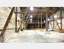 Großer, offener Innenraum einer Scheune oder Lagerhalle mit sichtbaren Holzbalken, Backsteinwänden und einem offenen Holzdach. Der Raum hat einen Betonboden, hölzerne Stützstrukturen und vereinzelte Leuchten an der Decke.