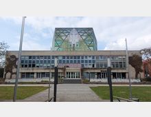 Modernes Gebäude mit einer rechteckigen Betonfassade und einem gläsernen Aufbau in geometrischem Design. Davor führen ein gepflasterter Weg und Treppenstufen zum Eingangsbereich; umgeben von Rasenflächen und kahlen Bäumen.