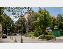 Straßenszene mit Kopfsteinpflaster, umgeben von dicht bepflanzten Bäumen und Wohnhäusern im Hintergrund. Im Vordergrund steht ein Schild mit der Aufschrift „Siemensstraße“ sowie ein historisches, grün lackiertes, rundes Toilettenhäuschen.