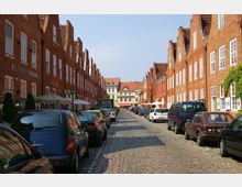 Straßenansicht einer gepflasterten Straße mit rechts und links zueinander symmetrischen roten Backsteinhäusern mit weißen Fenstern und Satteldächern. Geparkte Autos säumen die Straße, und im Hintergrund ist ein helles Gebäude mit einem roten Dach zu sehen.