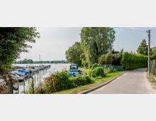 Teil eines Ufers mit einem schmalen Weg neben einem Zaun und grüner Vegetation, im Hintergrund eine Anlegestelle mit Booten auf einem ruhigen Fluss. Große Bäume und ein bewachsener Uferbereich prägen die Umgebung.