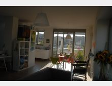 Helle Wohnung mit offenem Wohn- und Essbereich, moderner Einrichtung und großen Glasfronten, die auf einen Balkon mit Pflanzen und Stadtblick führen. Im Innenraum sind ein Esstisch, Regale, eine Küchenzeile und Dekorationen wie Sonnenblumen sichtbar.