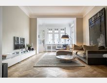 Wohnzimmer in modernem Stil mit hellem Parkettboden, einer beigen Wand mit Flachbildfernseher, einem großen beigen Sofa, einer Stehlampe und einem runden Couchtisch. Der Raum ist durch große Fenster mit weißen Rahmen lichtdurchflutet, und im Hintergrund ist ein Schreibtischbereich sichtbar.