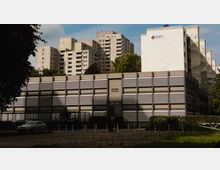 Mehrstöckige Wohngebäude aus Beton im Hintergrund, mit einem modernen Flachbau im Vordergrund, der eine Fassade aus grauen Paneelen und horizontalen weißen Lamellen hat. Links steht ein geparktes Auto, und im Vordergrund befindet sich eine asphaltierte Fläche mit vereinzeltem Grasbewuchs.