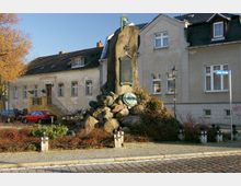 Denkmal aus großen Steinen mit einer Gedenktafel und einer Büste an der Spitze, umgeben von niedrigen Pflanzen und Kettenpfosten. Im Hintergrund stehen Wohnhäuser mit verputzten Fassaden an einer gepflasterten Straße, ein Straßenschild mit der Aufschrift „Breite Straße“ ist sichtbar.