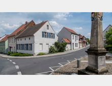 Straßenszene in einer Kleinstadt mit traditionellen Wohnhäusern, weißer Fassade und roten Ziegeldächern. Im Vordergrund steht ein historischer Wegweiser aus Stein mit Inschriften und Wappen, umgeben von gepflastertem Gehweg.