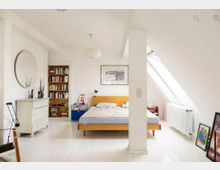 Minimalistisch eingerichtetes Schlafzimmer in einer Dachgeschosswohnung mit weiß gestrichenen Wänden und Holzmöbeln. Im Raum befinden sich ein Holzbett, ein Bücherregal, eine Kommode mit Spiegel und dekorativen Pflanzen sowie ein schräges Dachfenster, das natürliches Licht hereinlässt.