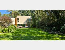 Modernes Wohnhaus mit flachen, hellen Fassaden und großen Fenstern, umgeben von einem gepflegten Garten mit Rasenfläche, Bäumen und dichten Sträuchern. Im Vordergrund befinden sich Sitzmöbel auf einer Terrasse, die an den Garten grenzt.