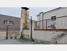 Dachterrasse mit einem Geländer aus Metall und weiß gekachelten Wänden, teilweise mit Graffiti besprüht. Im Hintergrund ist ein älteres Gebäude mit rotem Ziegeldach und Dachfenster zu sehen.