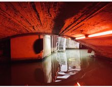 Backstein-Gewölbe eines unterirdischen Kanals oder Tunnels, teilweise in rotes Licht getaucht, mit stehendem Wasser am Boden und einer Metalltreppe, die zu einer Öffnung nach oben führt.