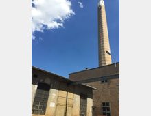Industrielle Gebäudefassade mit großen Fenstern und einer Metalltür, daneben ein hoher, runder Schornstein aus Ziegeln vor blauem Himmel mit vereinzelten Wolken.
