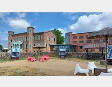 Historisches Backsteingebäude mit grünen Fensterrahmen und Türmen, kombiniert mit einem modernen Anbau aus Glas. Im Vordergrund ein sandiger Bereich mit Strandkörben, Sonnenschirmen aus Stroh und bunten Sitzmöbeln, umgeben von einer niedrigen Steinmauer.