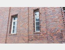 Backsteinfassade mit zwei schmalen, sprossengeteilten Fenstern, deren Umrahmung durch hervorgehobene Ziegelsteine akzentuiert ist. Die Backsteine variieren in Rottönen und zeigen eine strukturierte, historische Bauweise.