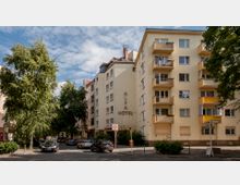 Straßenszene mit mehrstöckigen Wohngebäuden, darunter ein Hotel mit der Aufschrift &quot;ALFA HOTEL&quot; auf der Fassade. Die Umgebung ist von Bäumen gesäumt, es sind parkende Autos und ein Schild für eine Tempo-30-Zone sichtbar.