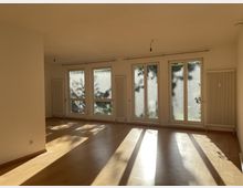 Leerer Raum mit hellen Holzböden, drei großen bodentiefen Fenstern mit Blick ins Grüne, zwei Heizkörpern an den Wänden und einfallendem Sonnenlicht.