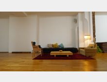 Wohnzimmer mit Holzboden und minimalistischer Einrichtung; zentral steht ein niedriger Holztisch auf einem roten Teppich, umgeben von einem schwarzen Sofa, einem blauen Sessel und einem beigen Sessel. Im Hintergrund befinden sich eine weiße Wand, ein Heizkörper, eine Stehlampe und gestapelte Yogamatten.