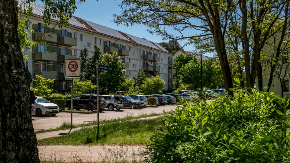 Blick auf eine Wohnsiedlung mit mehrstöckigen Mehrfamilienhäusern und Balkonen, davor ein Parkplatz mit zahlreichen Autos entlang einer Straße. Im Vordergrund stehen Bäume, Sträucher und eine Grünfläche; ein Verkehrsschild weist auf eine Tempo-30-Zone hin.