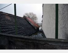 Blick auf eine Dachfläche mit dunklen Ziegeln und einem kleineren Bereich aus roten Ziegeln, flankiert von einer verputzten Hauswand rechts. Im Hintergrund ist in einer nebligen Landschaft ein kahler Baum und ein Gewässer erkennbar.