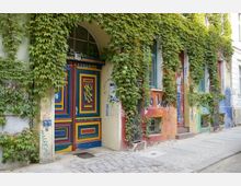 Fassade eines Altbaus mit buntem, künstlerisch bemaltem Eingangstor und umliegender Wand, die von dichtem grünen Efeu überwuchert ist. Der Gehweg davor besteht aus Kopfsteinpflaster, und die Wände sind mit Graffiti verziert.
