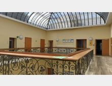 Innenansicht eines Gebäudes mit einem Dach aus gewölbtem, gerastertem Glas, das Tageslicht hereinlässt. Der Raum hat gelbe Wände, eine umlaufende Galerie mit kunstvollen schmiedeeisernen Geländern und Türen, die in verschiedene Räume führen.