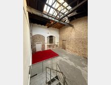 Innenraum einer ehemaligen Industriehalle mit hohen Ziegelwänden, sichtbaren Stahlträgern und großem Oberlicht im Dach. Unten ein Betonboden mit roter Plattform sowie eine Metalltreppe und ein weißer Wandbereich mit Rundbögen und einem vergitterten Fenster.