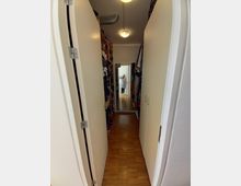 Ein schmaler Flur oder begehbarer Kleiderschrank mit Parkettboden, hohen Regalen voller Kleidung an beiden Seiten und einer Spiegeltür am Ende des Gangs. Zwei Deckenlampen beleuchten den Raum, und eine Person ist im Spiegel zu sehen.